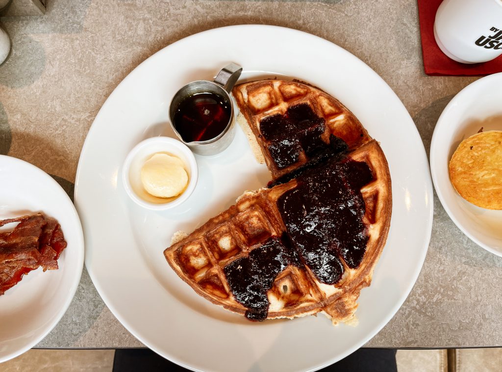 PLate of waffles tooped with blueberry compote, side orders of bacon on one side and hashbrown on the other side.